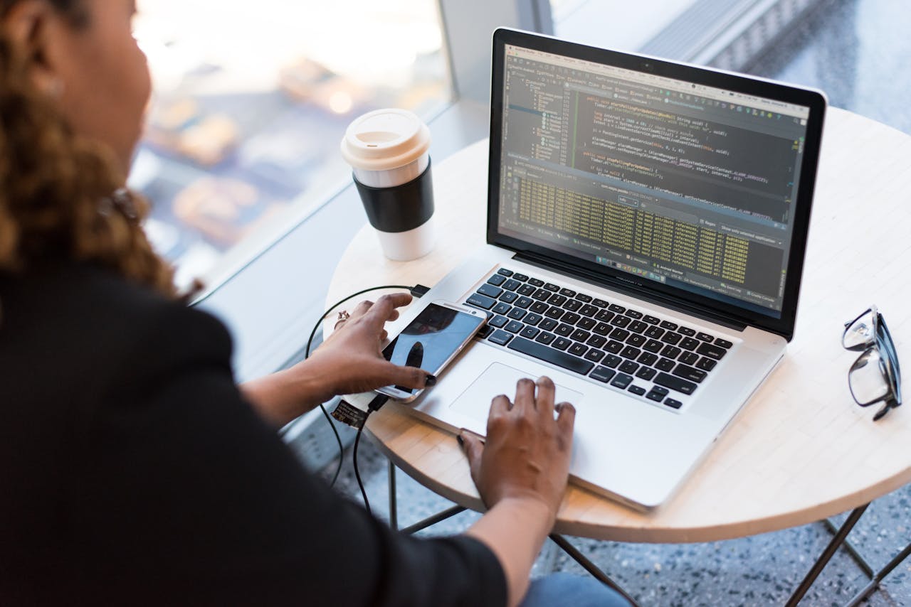 journey Black woman programming on a laptop with coffee, smartphone, and glasses on a desk in an office.