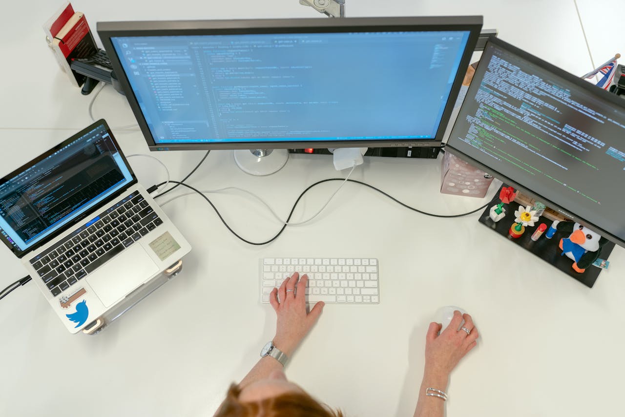 who-we-are A female software engineer coding on dual monitors and a laptop in an office setting.